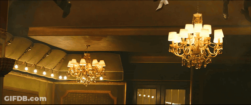 A Man In A Red And Gold Spiderman Suit Stands In Front Of A Chandelier GIF