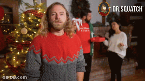 A Man In A Red And Gray Sweater Is Standing In Front Of A Christmas Tree GIF