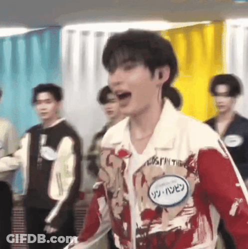 A Man In A Red And White Jacket With A Name Tag On His Chest Is Standing In Front Of A Group Of People . GIF