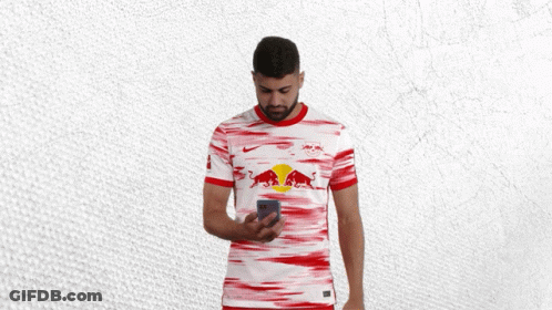 A Man In A Red And White Jersey With A Bull On It Looks At His Phone GIF