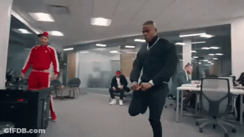 A Man In A Red And White Tracksuit Is Stretching His Legs In An Office While Other Men Sit At Desks . GIF