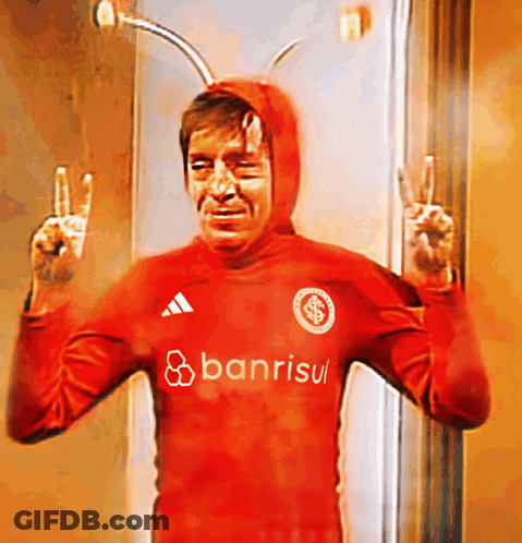 A Man In A Red Banrisul Shirt Giving The Peace Sign GIF
