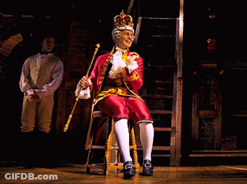 A Man In A Red Costume With A Crown On His Head Is Sitting On A Stool GIF