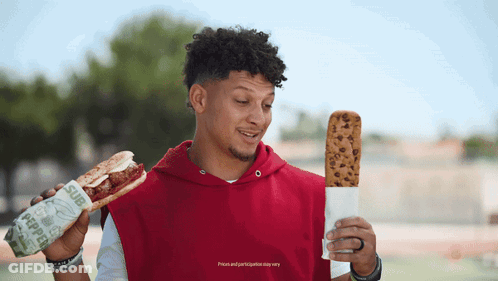 A Man In A Red Hoodie Is Holding A Sub Sandwich And A Cookie GIF