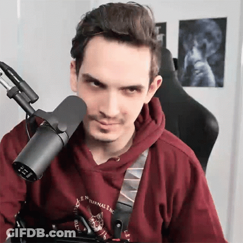 A Man In A Red Hoodie Is Sitting In Front Of A Microphone With A Picture Of A Skull On The Wall Behind Him GIF