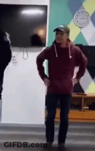A Man In A Red Hoodie Is Standing In Front Of A Clock On The Wall . GIF