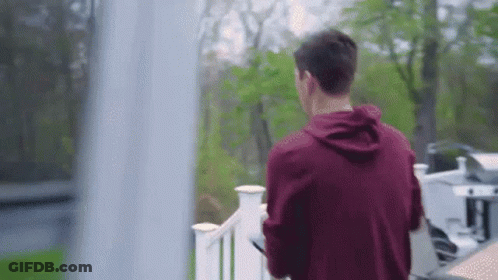 A Man In A Red Hoodie Is Standing On A Balcony Looking Out A Window . GIF