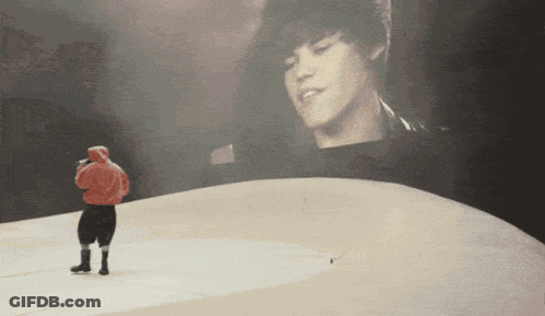 A Man In A Red Hoodie Stands In Front Of A Large Screen With A Picture Of Justin Bieber GIF