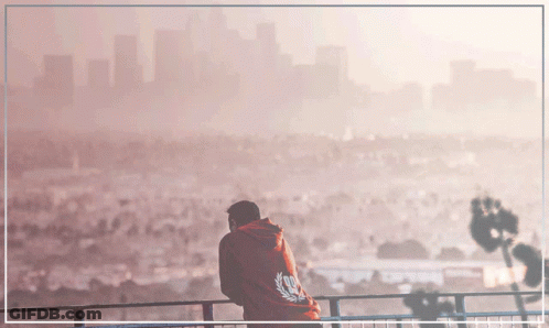A Man In A Red Hoodie Stands On A Balcony With A City In The Background GIF