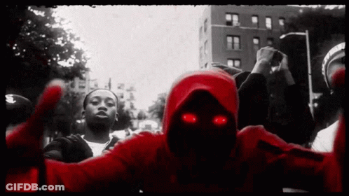 A Man In A Red Hoodie With Red Eyes Stands In A Crowd Of People GIF