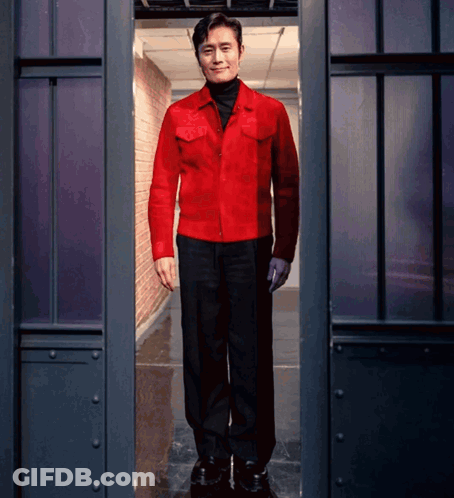 A Man In A Red Jacket And Black Pants Stands In A Dark Room GIF
