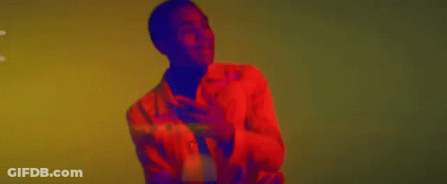 A Man In A Red Jacket And Black Turtleneck Is Dancing In Front Of A Yellow Wall . GIF