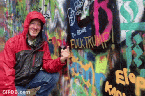 A Man In A Red Jacket Giving A Thumbs Up In Front Of A Wall With Graffiti On It That Says Fuck Trump GIF