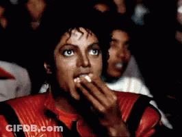 A Man In A Red Jacket Is Eating Something While Watching A Movie GIF