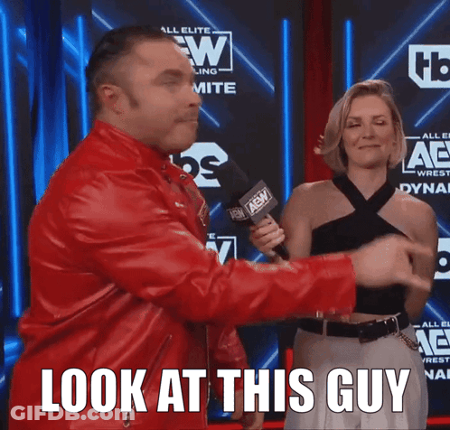 A Man In A Red Jacket Is Holding A Microphone And A Woman Behind Him Says " Look At This Guy " GIF