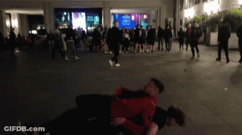 A Man In A Red Jacket Is Holding Another Man In A Crowd GIF