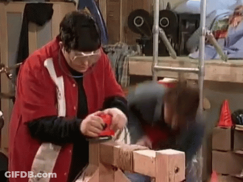 A Man In A Red Jacket Is Sanding A Piece Of Wood While A Woman Watches . GIF