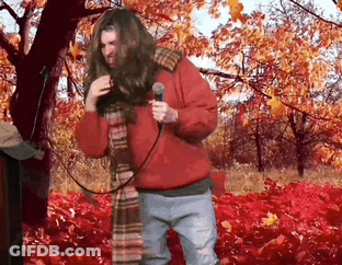 A Man In A Red Jacket Is Singing Into A Microphone In A Field Of Leaves . GIF
