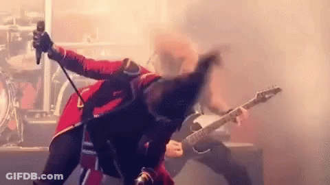 A Man In A Red Jacket Is Singing Into A Microphone While Another Man Plays A Guitar . GIF