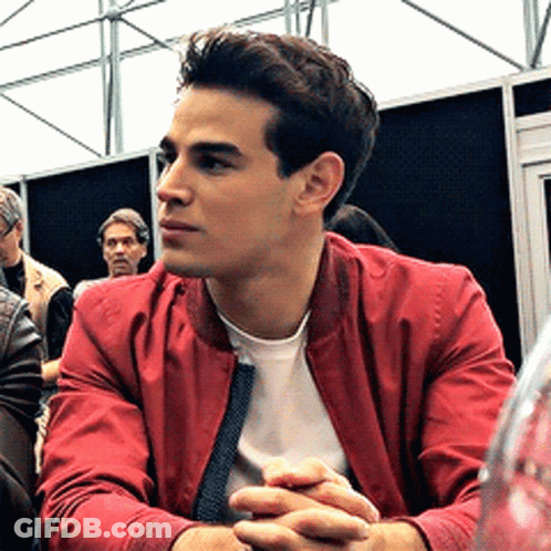 A Man In A Red Jacket Is Sitting At A Table With His Hands Folded . GIF