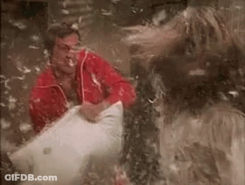 A Man In A Red Jacket Is Sitting In A Pile Of Feathers While A Woman Throws Feathers At Him . GIF