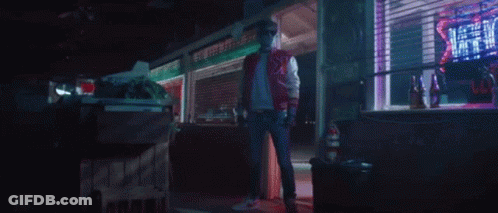 A Man In A Red Jacket Is Standing In Front Of A Store With A Neon Sign That Says ' Hot ' On It . GIF