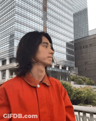 A Man In A Red Jacket Is Standing In Front Of A Tall Building GIF