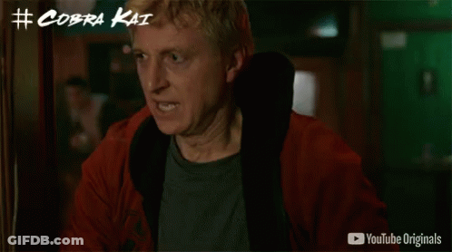 A Man In A Red Jacket Is Standing In Front Of A Wall With # Cobra Kai Written Above Him GIF
