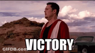 A Man In A Red Jacket Is Standing In The Desert With The Word Victory Written In White Letters . GIF