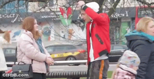 A Man In A Red Jacket Is Standing Next To A Woman In A Pink Jacket On A City Street . GIF
