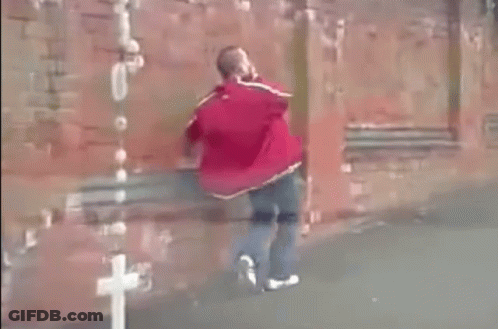 A Man In A Red Jacket Is Walking In Front Of A Red Brick Wall . GIF