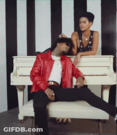 A Man In A Red Jacket Sits On A Bench Next To A Woman Leaning On A White Piano GIF