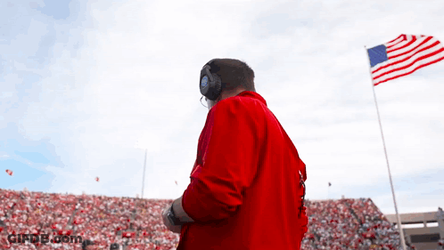 A Man In A Red Jacket Stands In Front Of A Large American Flag GIF