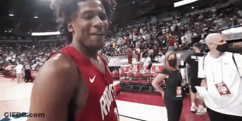 A Man In A Red Jersey Is Standing On A Basketball Court With A Crowd Behind Him . GIF