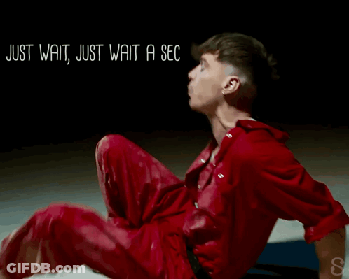 A Man In A Red Jumpsuit Is Sitting On The Floor With The Words Just Wait Just Wait A Sec Written Above Him GIF