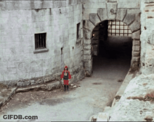 A Man In A Red Knight Costume Stands In Front Of A Stone Building GIF