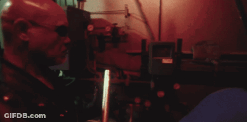 A Man In A Red Mask Is Standing In A Dark Room With A Red Light Behind Him GIF