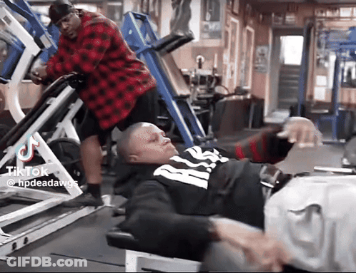 A Man In A Red Plaid Shirt Is Standing Next To A Man In A Black And White Shirt On A Leg Press Machine . GIF