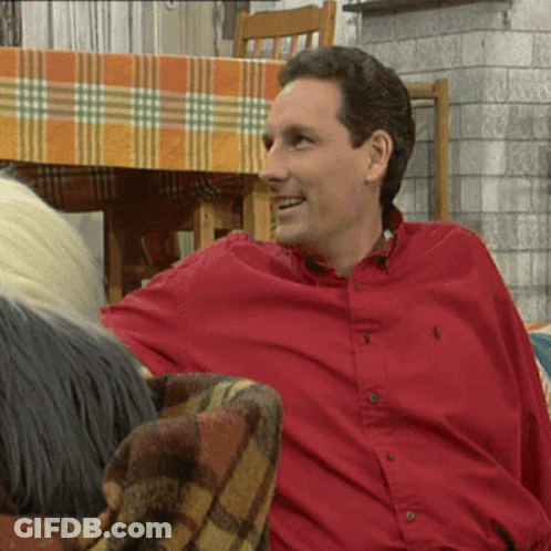 A Man In A Red Polo Shirt Smiles While Sitting On A Couch GIF