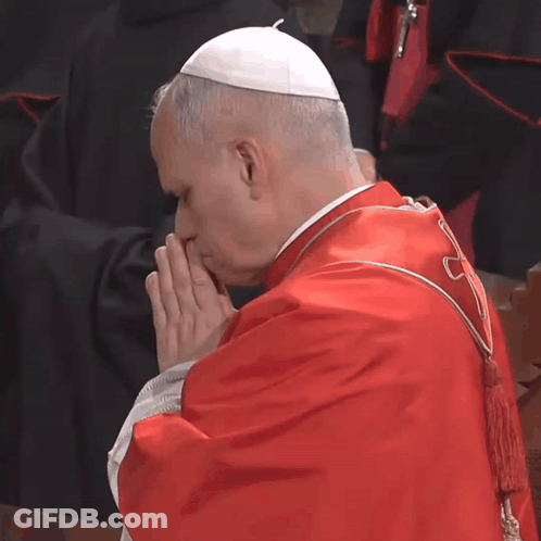 A Man In A Red Robe And White Hat Prays GIF