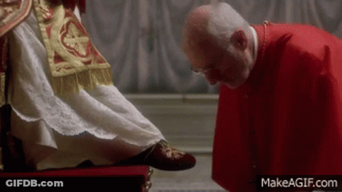 A Man In A Red Robe Is Kissing The Feet Of A Man In A White Dress . GIF