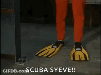 A Man In A Red Scuba Suit And Yellow Gloves Is Standing In A Room . GIF