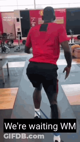 A Man In A Red Shirt And Black Shorts Is Running In A Gym With The Words `` We 'Re Waiting Wm '' . GIF