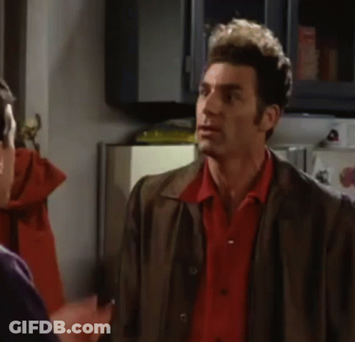 A Man In A Red Shirt And Brown Jacket Is Making A Funny Face While Talking To Another Man In A Kitchen . GIF