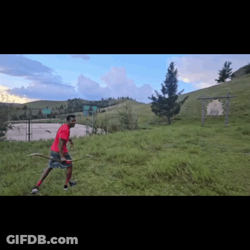A Man In A Red Shirt Holds A Bow And Arrow In A Field GIF