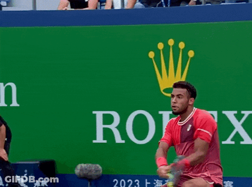 A Man In A Red Shirt Holds A Tennis Racquet In Front Of A Rolex Sign GIF