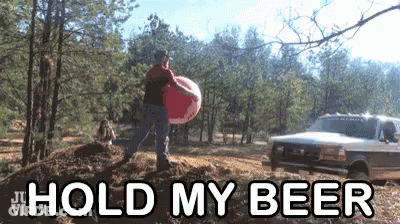 A Man In A Red Shirt Is Carrying A Large Inflatable Ball With The Words Hold My Beer Behind Him GIF