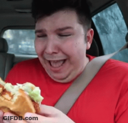 A Man In A Red Shirt Is Eating A Hamburger While Wearing A Seat Belt GIF