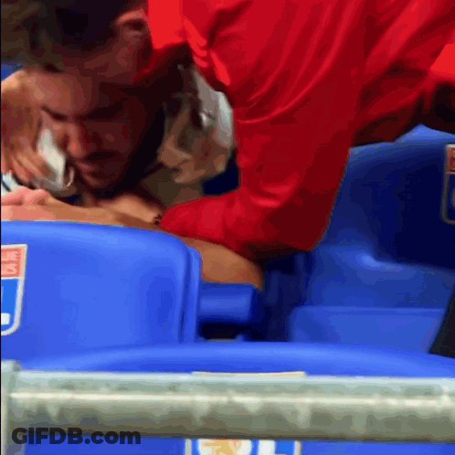 A Man In A Red Shirt Is Helping Another Man In A Stadium Seat GIF