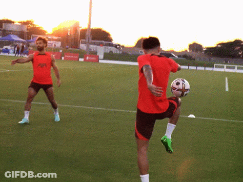 A Man In A Red Shirt Is Kicking A Soccer Ball While Another Man Looks On GIF